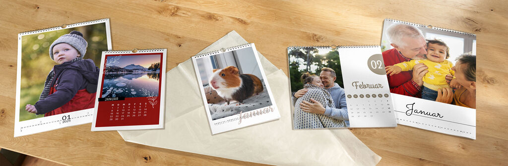 Drie kalenders liggen op een tafel naast kerstversiering zoals inpakpapier en een lichtslinger.